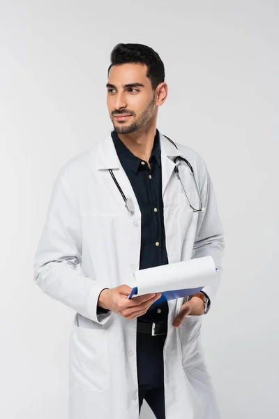 Muslim doctor in white coat holding clipboard and looking away isolated on grey — Stock Photo