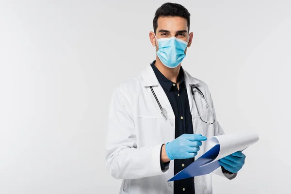 Joven médico árabe en máscara médica sujetando el portapapeles y mirando a la cámara aislada en gris - foto de stock