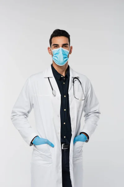 Médico árabe en máscara médica y guantes de látex mirando a la cámara aislada en gris - foto de stock