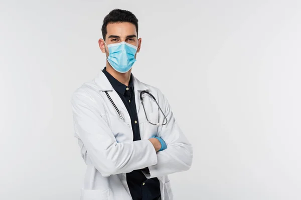 Joven médico musulmán en máscara médica y guante de látex mirando a la cámara aislada en gris - foto de stock