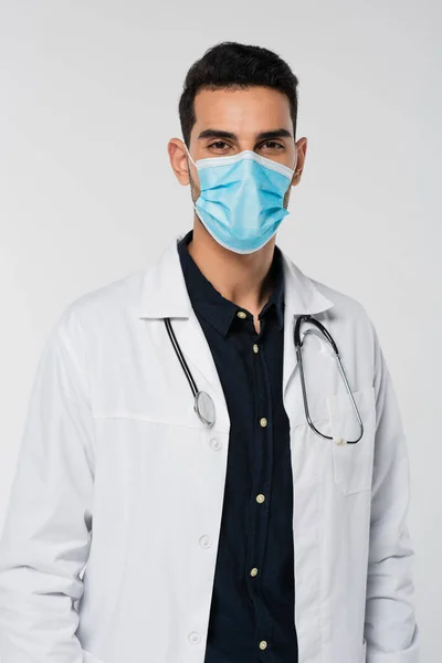 Médico árabe en máscara médica mirando la cámara aislada en gris - foto de stock