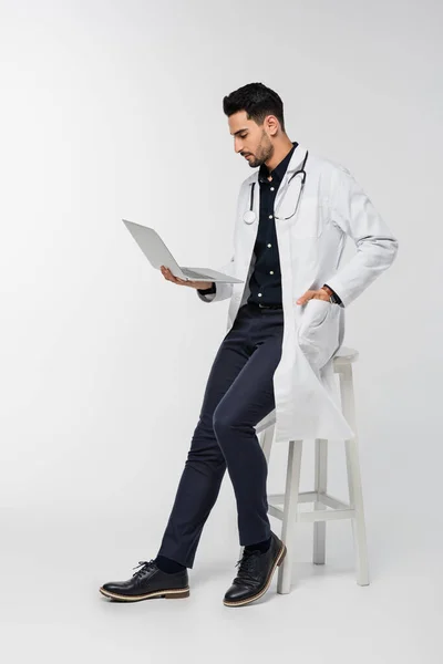 Vista lateral do médico árabe usando laptop perto da cadeira no fundo cinza — Fotografia de Stock