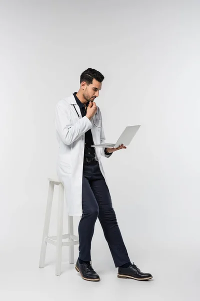 Side view of muslim doctor using laptop near chair on grey background — Stock Photo