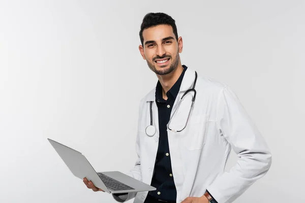 Allegro medico arabo che tiene il computer portatile e guarda la fotocamera isolata sul grigio — Foto stock