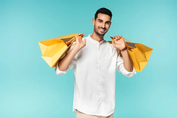 Hombre árabe sonriente sosteniendo bolsas de compras y mirando a la cámara aislada en azul - foto de stock