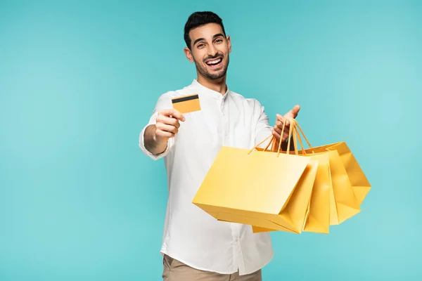 Позитивний арабський чоловік тримає кредитну картку і жовті сумки для покупок ізольовані на синьому — стокове фото