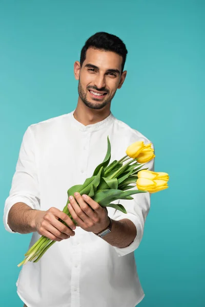 Желтые тюльпаны в руках улыбающегося арабского человека на размытом фоне, изолированном на голубом — стоковое фото