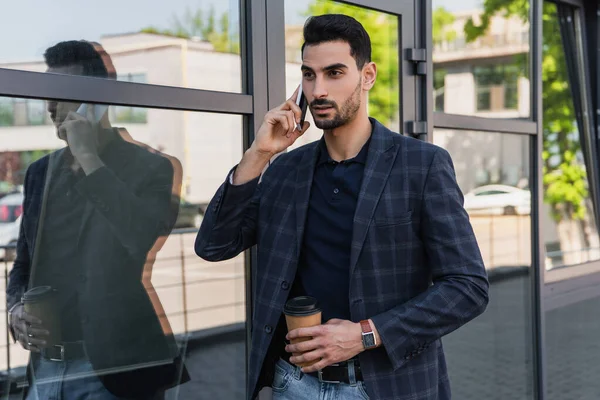Hombre de negocios árabe hablando por teléfono móvil y sosteniendo una taza de papel cerca del edificio - foto de stock