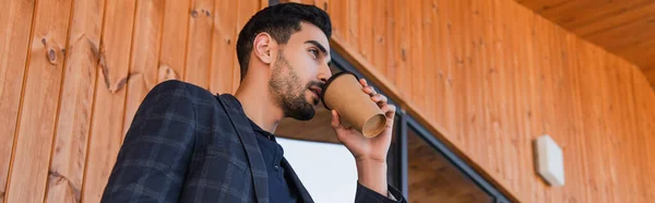 Vista de bajo ángulo del empresario árabe bebiendo café cerca del edificio con fachada de madera, pancarta - foto de stock