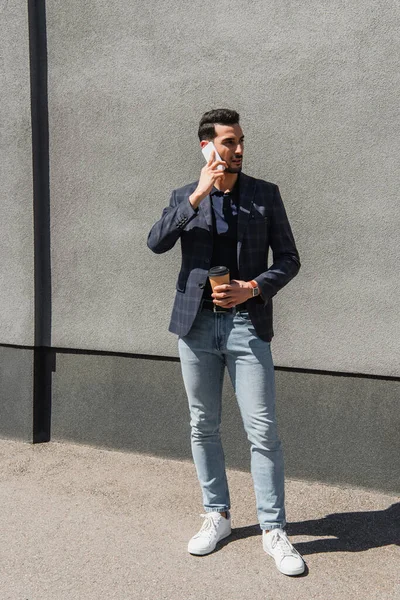 Arabian businessman talking on smartphone while holding coffee to go near building — Stock Photo