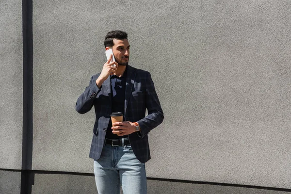 Hombre musulmán con chaqueta sosteniendo una taza de papel y hablando en un teléfono inteligente cerca de la pared de hormigón - foto de stock