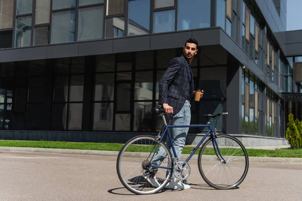 Joven gerente árabe sosteniendo una taza para llevar cerca de la bicicleta en la calle urbana - foto de stock