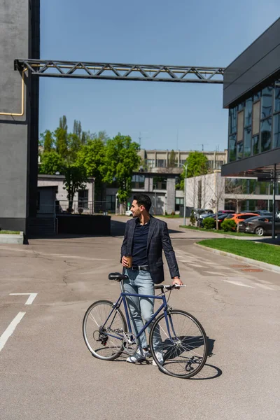 Vista lateral de hombre de negocios musulmán sonriente con bicicleta y café para ir de pie en la calle - foto de stock