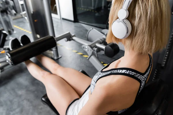Jovem mulher em fones de ouvido sem fio treinamento na máquina de exercício de extensão de perna — Fotografia de Stock