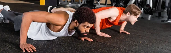 Hombres interracial en ropa deportiva haciendo flexiones en el centro deportivo, pancarta - foto de stock