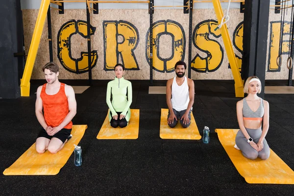 Interracial people sitting on fitness mats in thunderbolt pose near sports bottles — Stock Photo