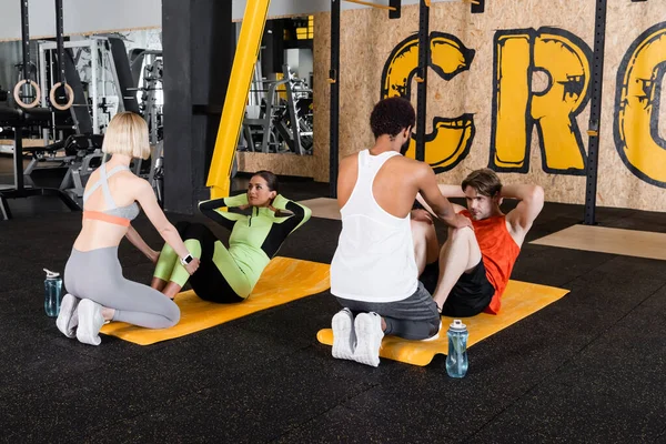 Entrenadores interraciales que apoyan a los jóvenes haciendo abdominales en el gimnasio - foto de stock