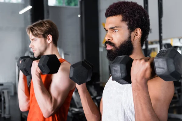 Bärtiger afrikanisch-amerikanischer Mann trainiert mit Hanteln in der Nähe eines verschwommenen Freundes — Stockfoto