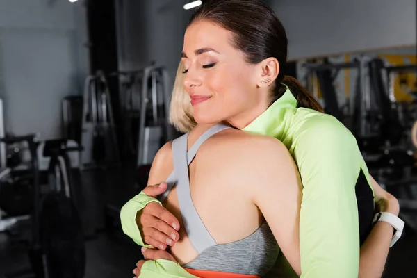 Happy trainer with closed eyes embracing sportswoman in gym — Stock Photo