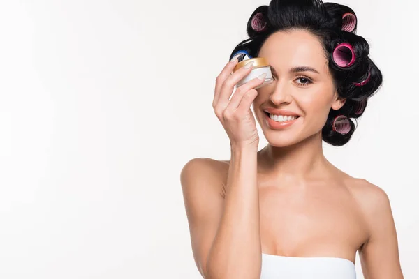 Sorrindo jovem morena com encrespadores no cabelo segurando recipiente creme perto do rosto isolado no branco — Fotografia de Stock