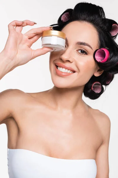 Sorridente giovane donna bruna con bigodini sui capelli che tengono il contenitore di crema vicino al viso isolato sul bianco — Foto stock