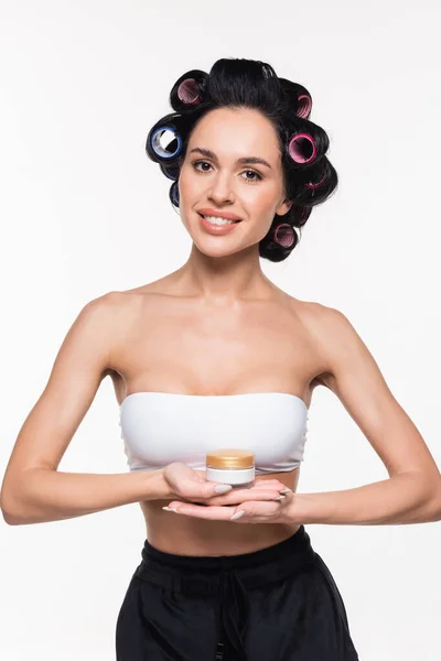 Lächelnde junge Frau in weißem BH mit Lockenwicklern auf dem Haar, die Cremebehälter in den Händen halten, isoliert auf weißem — Stockfoto