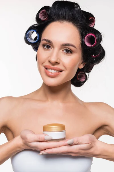 Portrait of smiling young woman in white bra with curlers on hair holding cream container in hands isolated on white — Stock Photo