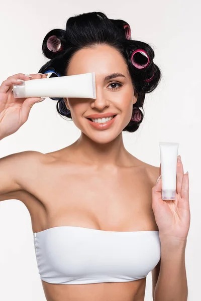 Sorrindo jovem mulher em rolos segurando tubos de creme na mão e perto do rosto isolado no branco — Fotografia de Stock