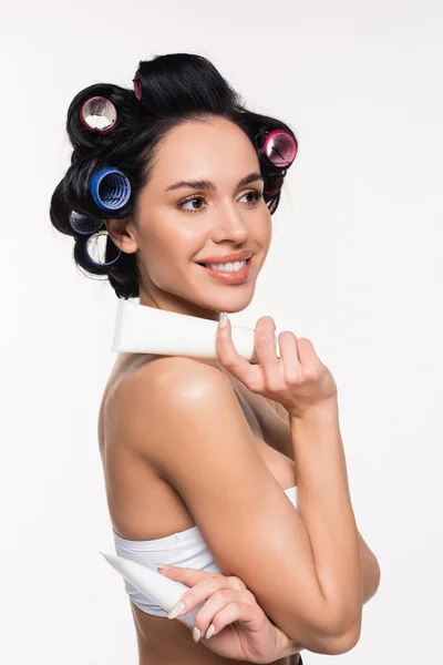 Smiling young woman in curlers holding cream tubes isolated on white — Stock Photo