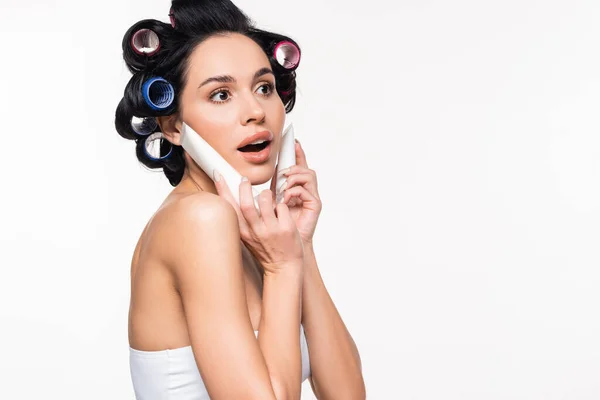 Animado jovem mulher com encrespadores na cabeça segurando tubos de creme perto do rosto com a boca aberta isolada no branco — Fotografia de Stock