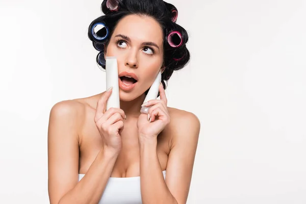 Excited young woman in top and curlers holding cream tubes near face with open mouth isolated on white — Stock Photo