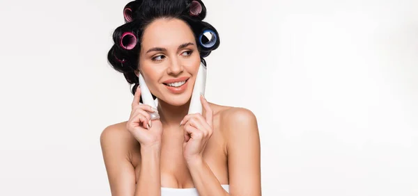 Smiling young woman in top and curlers holding cream tubes near face isolated on white, banner — Stock Photo