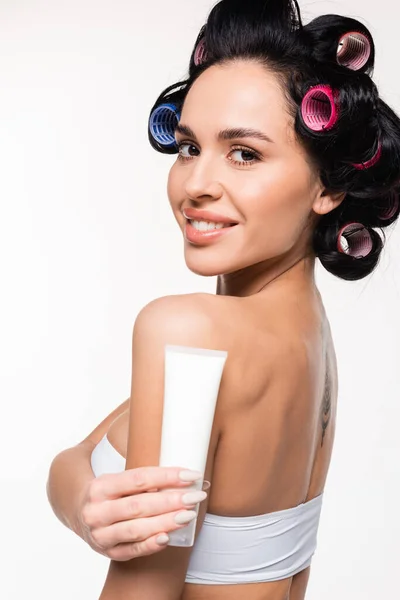 Cheerful young woman in curlers and top holding cream tube near shoulder isolated on white — Stock Photo