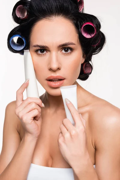 Ritratto di giovane donna con bigodini sulla testa tenendo tubi crema vicino al viso isolato su bianco — Foto stock