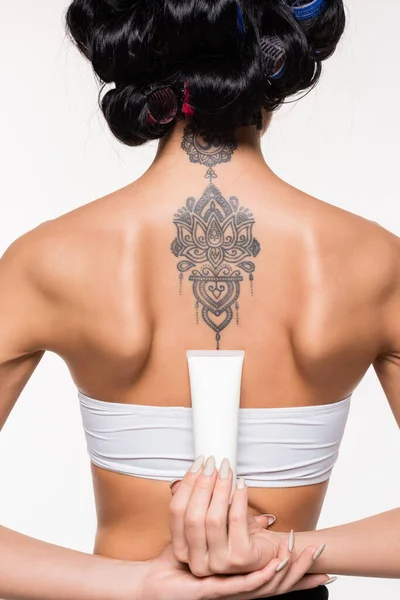 Vista posteriore di giovane donna in bigodini con tatuaggio tenuta tubo crema isolato su bianco — Foto stock