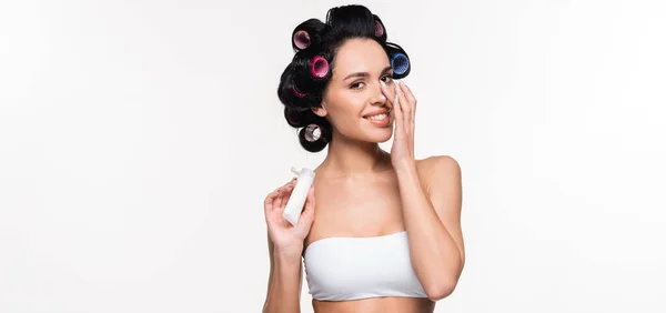 Sorrindo jovem mulher em rolos e sutiã aplicando creme no nariz e segurando tubo na mão isolado em branco, banner — Fotografia de Stock