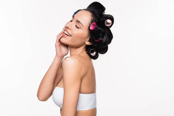 Jeune femme souriante en bigoudis et haut posant avec crème appliquée sur l'épaule, les yeux fermés et la main près du visage isolé sur blanc — Photo de stock