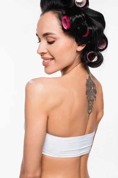 Усміхнена молода жінка в бігунках і татуювання на спині стоїть з прикладеним кремом на плечі ізольовано на білому — стокове фото