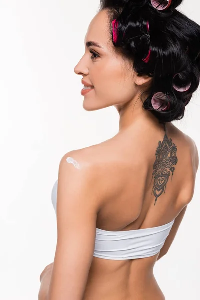 Sonriente mujer joven en rulos y tatuaje en la espalda de pie con crema aplicada en el hombro aislado en blanco - foto de stock