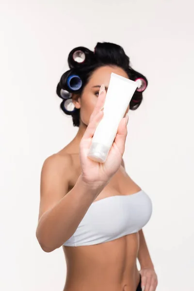 Jeune femme en bigoudis couvrant le visage avec tube à crème à la main isolé sur blanc — Photo de stock