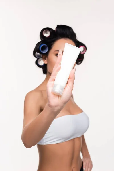 Young woman in curlers covering face with cream tube in hand isolated on white — Stock Photo