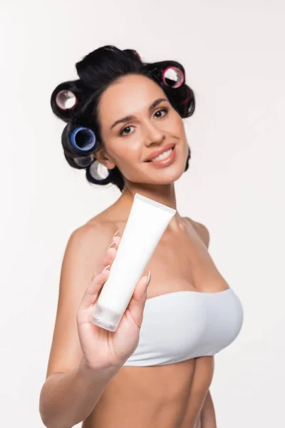 Sonriente mujer joven en rulos y sujetador mostrando tubo de crema aislado en blanco - foto de stock