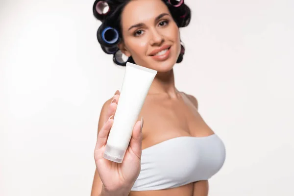 Sorridente giovane donna in bigodini e top mostrando tubo crema e guardando la fotocamera isolata su bianco — Foto stock
