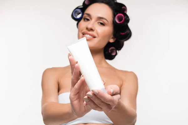Joyeuse jeune femme en bigoudis et haut montrant tube de crème isolé sur blanc — Photo de stock
