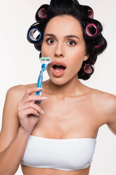 Amazed young woman in curlers holding razor near face with open mouth isolated on white — Stock Photo