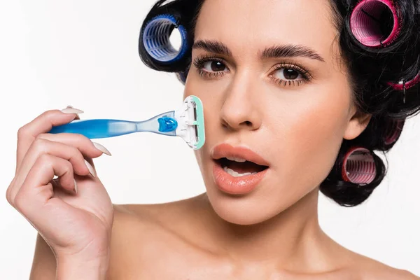 Retrato de mujer joven asombrada en rulos sosteniendo la navaja cerca de la cara con la boca abierta aislada en blanco - foto de stock