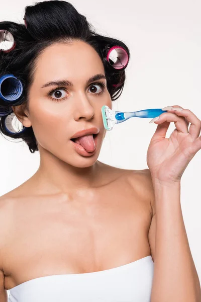 Young woman in curlers and top grimacing with sticking out tongue and holding razor near face isolated on white — Stock Photo