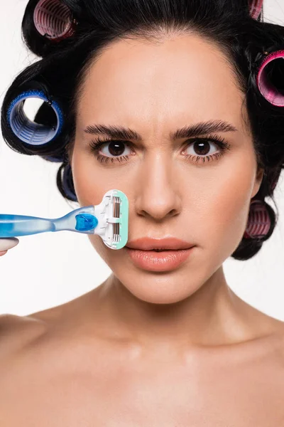 Retrato de mujer joven confundida en rulos sosteniendo afeitadora cerca de la cara aislada en blanco - foto de stock