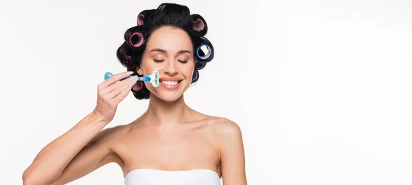 Cheerful young woman in curlers and top holding razor near face with closed eyes isolated on white, banner — Stock Photo