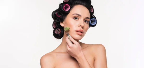 Grave jovem mulher em curlers fazendo massagem facial com rolo de jade isolado em branco, banner — Fotografia de Stock